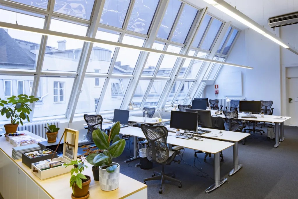 Ett kontor med stora lutande glasfönster, skrivbord, kontorsstolar och krukväxter.