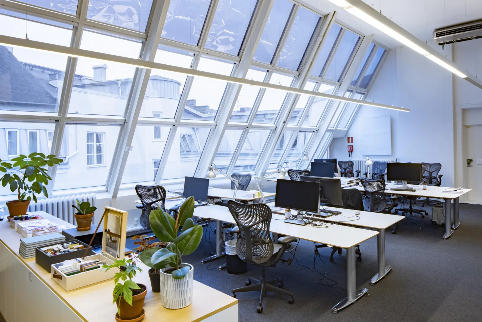 Ett kontor med stora lutande glasfönster, skrivbord, kontorsstolar och krukväxter.