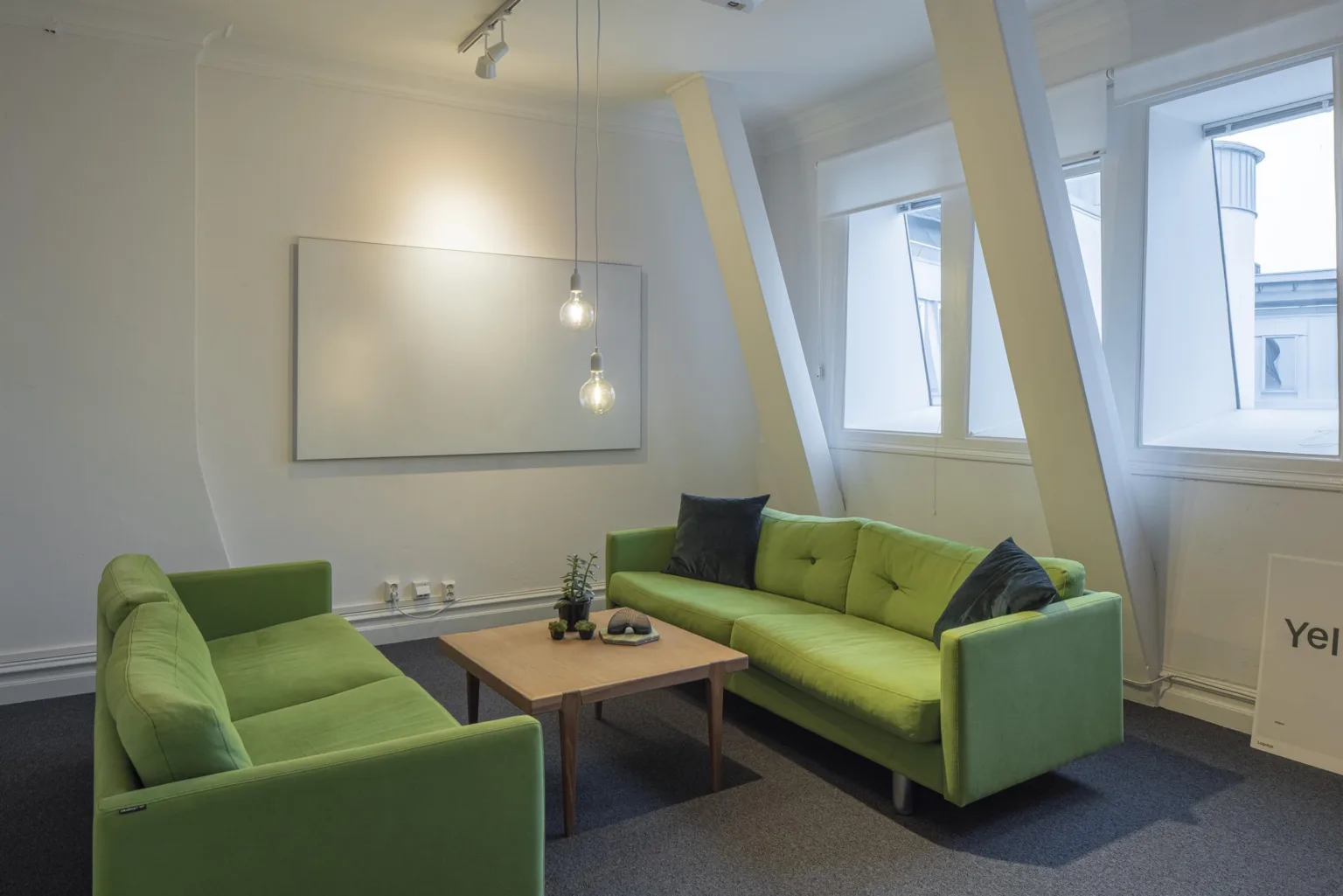 3. Ett vardagsrumsområde med två gröna soffor, ett träbord i mitten, en vit tavla på väggen och stora fönster som släpper in ljus.