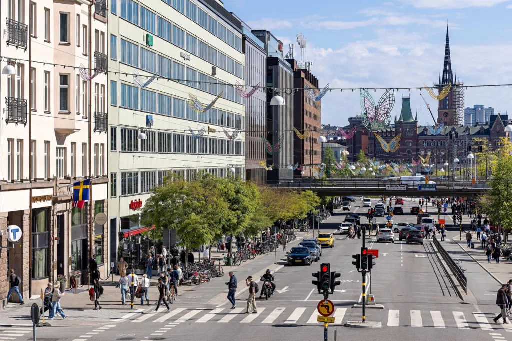 En stadsvy med en trafikerad gata, byggnader, fotgängare och en bro, under en klarblå himmel.