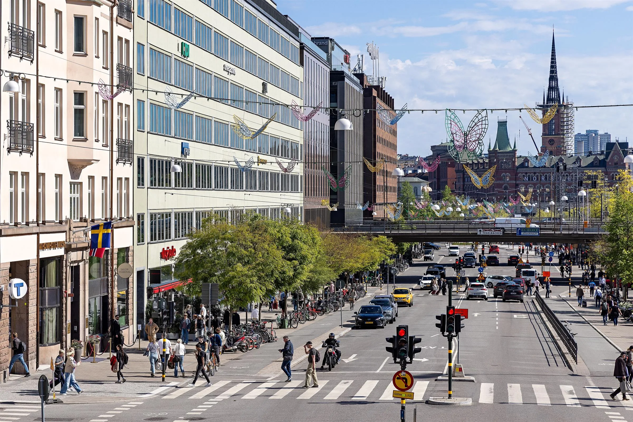 En stadsvy med en trafikerad gata, byggnader, fotgängare och en bro, under en klarblå himmel.