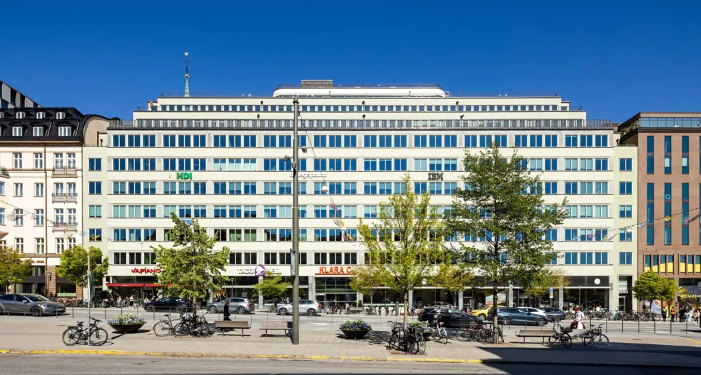 3. Fasad av en stor byggnad med glasfönster, träd och en gata med bänkar och parkeringsplatser.