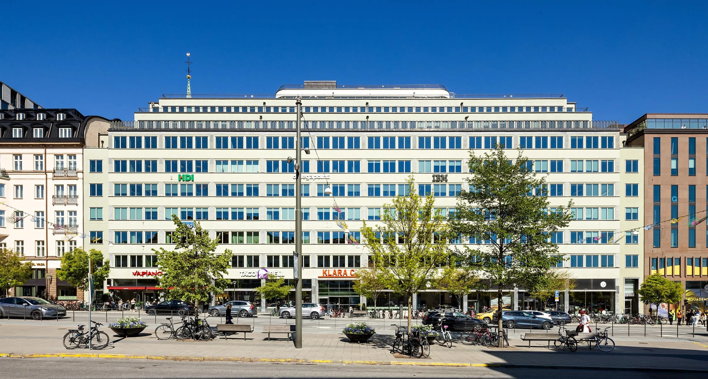 3. Fasad av en stor byggnad med glasfönster, träd och en gata med bänkar och parkeringsplatser.