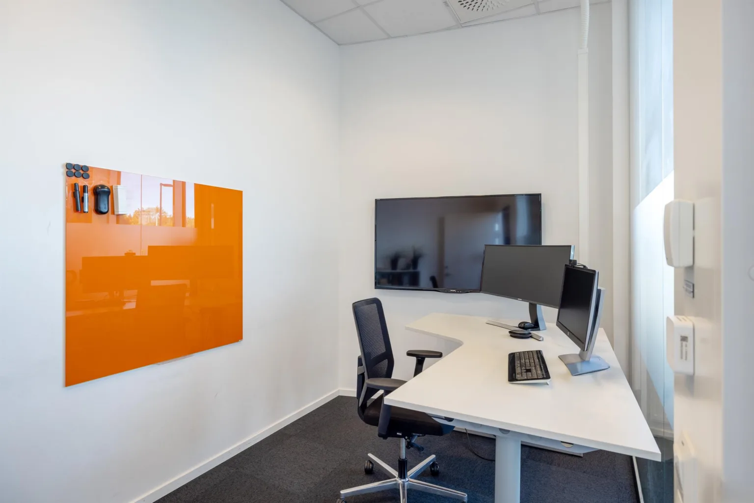 Ett litet kontorsrum med ett skrivbord, två datorskärmar, en svart stol och en orange anslagstavla på väggen.