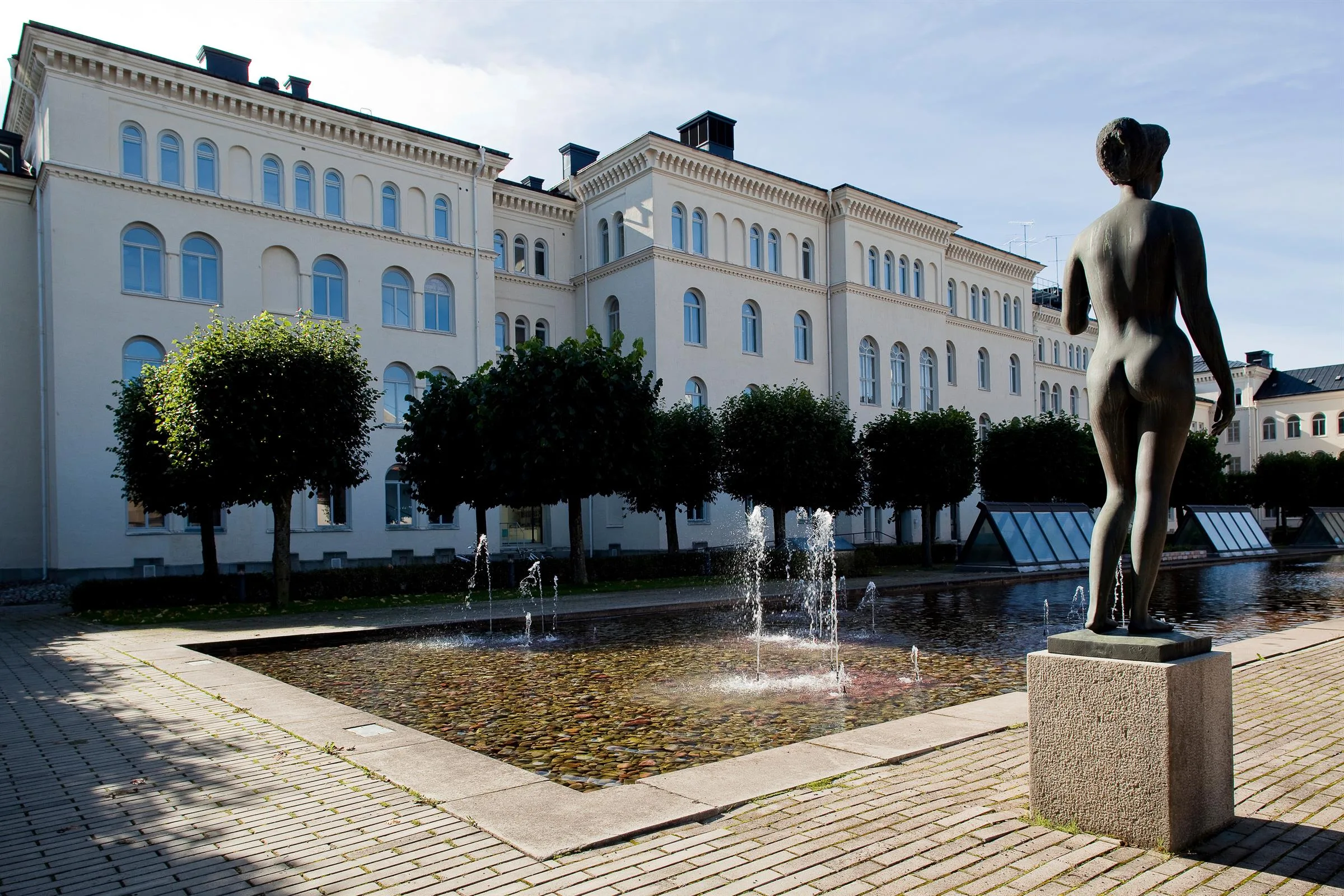 En fontän med vattenstrålar framför en vit historisk byggnad och välklippta träd.