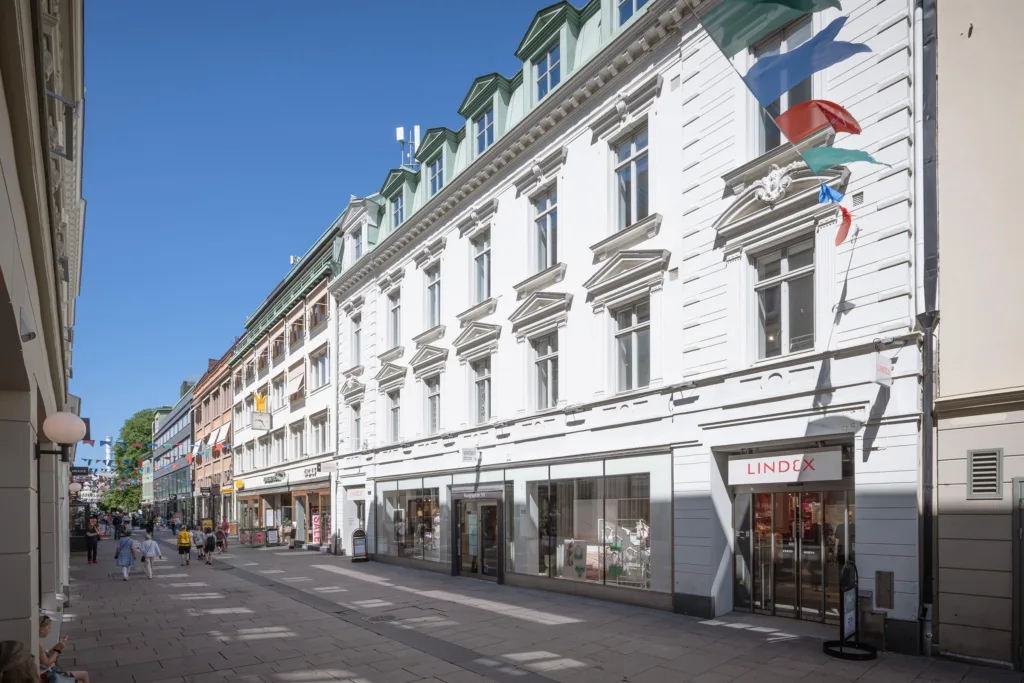 En lång vit byggnad med gröna tak och stora fönster, med butiker och trottoar längs fasaden.