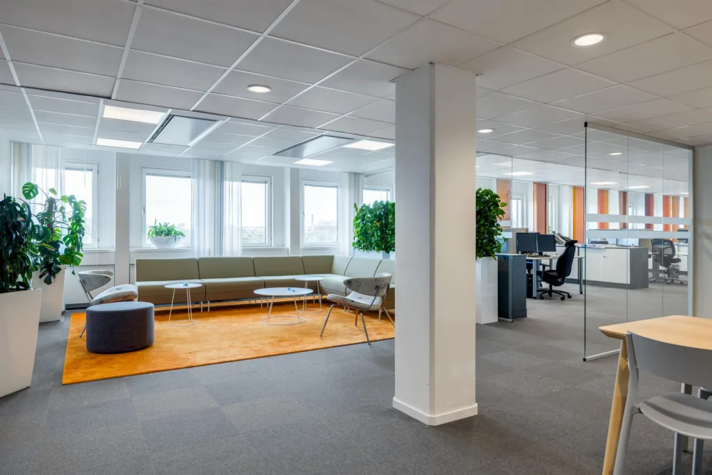 Ett loungeområde med soffor, ett orange mattområde och växter, bredvid ett öppet kontor med skrivbord.