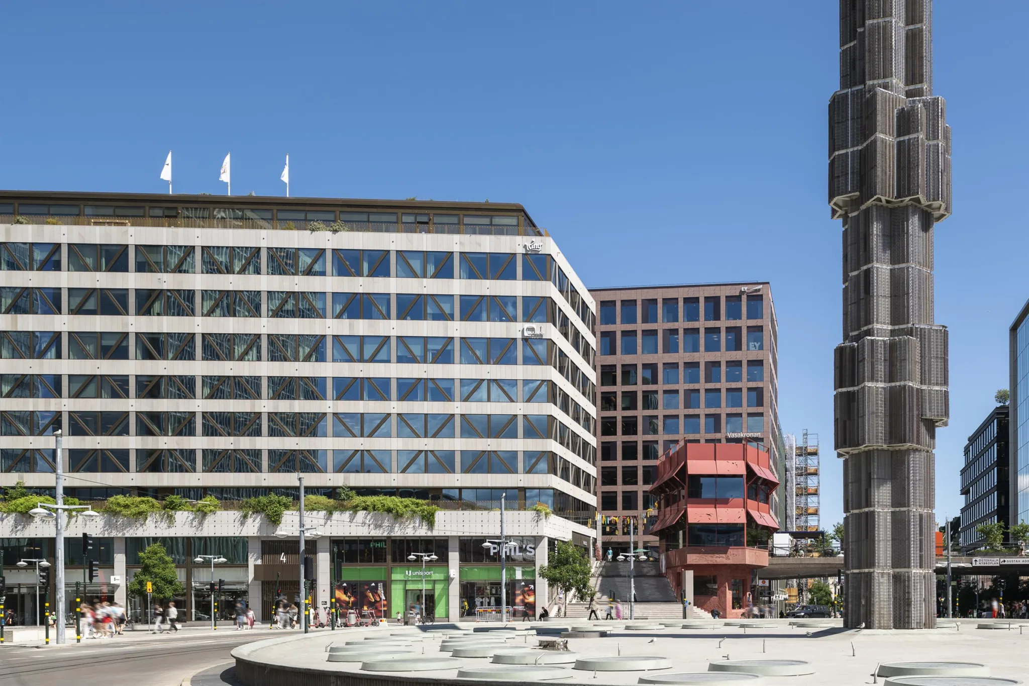 En exteriör av moderna byggnader med glasfasader, en fontän och en klar blå himmel.