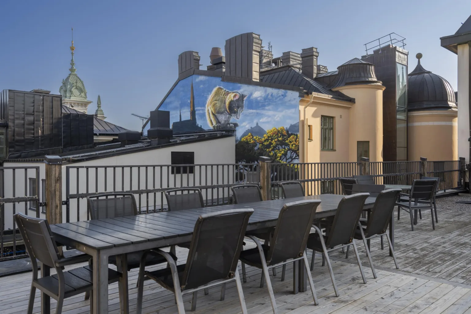 En takterrass med lång matbord, svarta stolar och utsikt över stadens byggnader och en muralmålning.