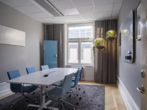 Ett mötesrum med ett vitt bord, blå stolar, en blå förvaringsskåp och hängande växter vid fönstret.