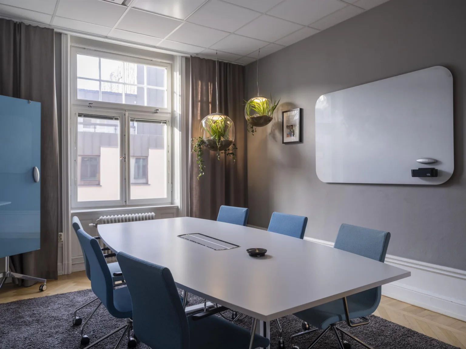 Ett konferensrum med ett stort vitt bord, blå stolar, en vit skrivtavla på väggen och stora fönster.