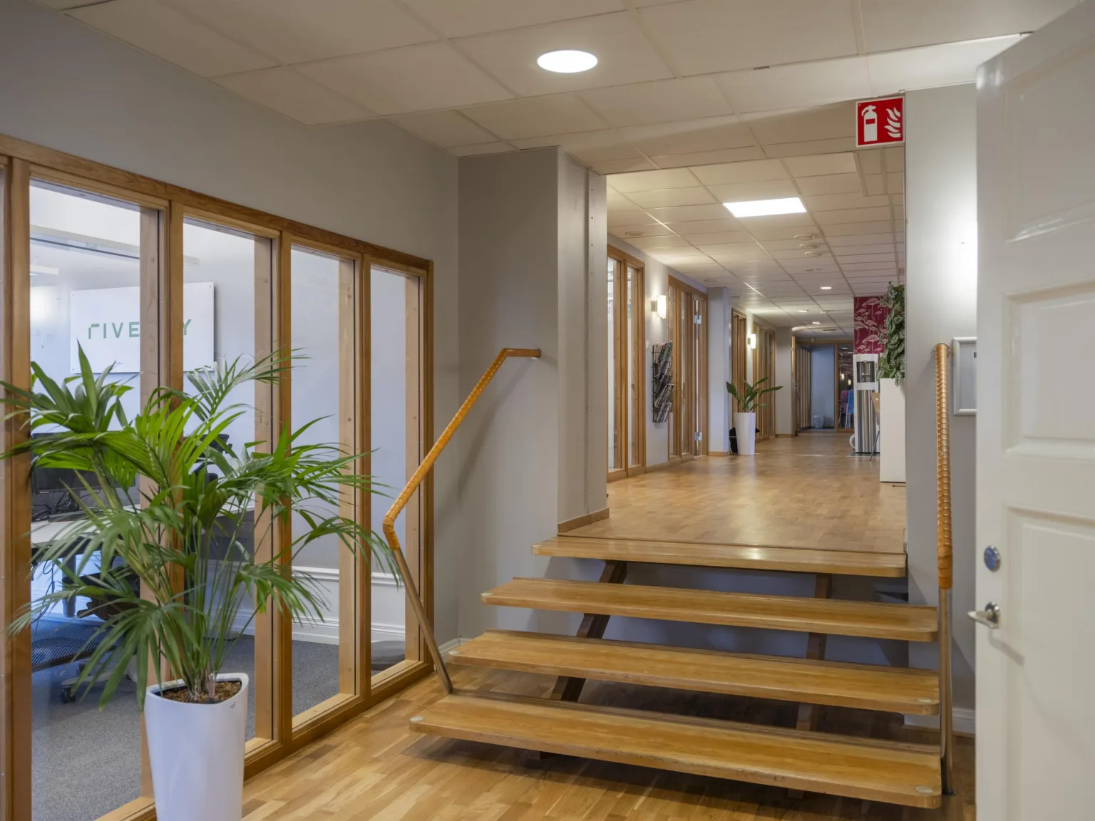 En kontorshall med trätrappor, glasväggar och gröna växter i vita krukor.