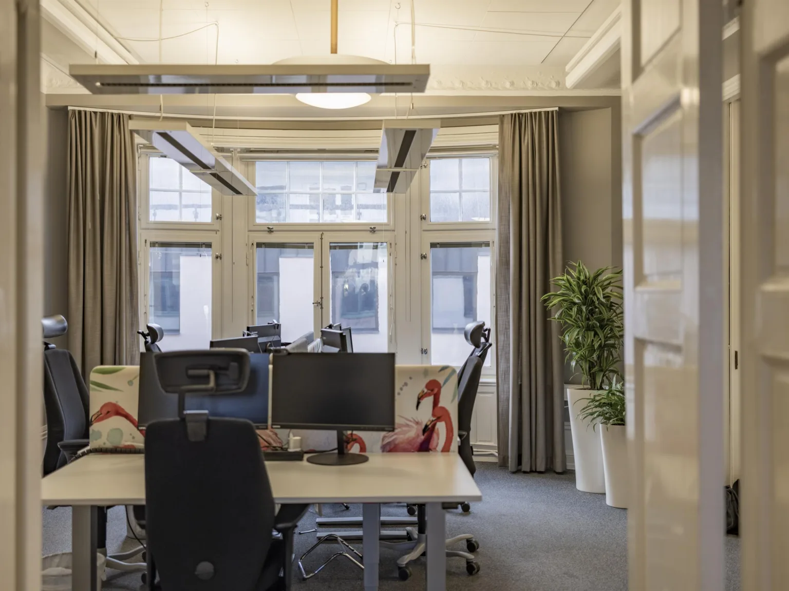 En kontorsmiljö med skrivbord, datorskärmar och stolar samt stora fönster och grå gardiner i bakgrunden.