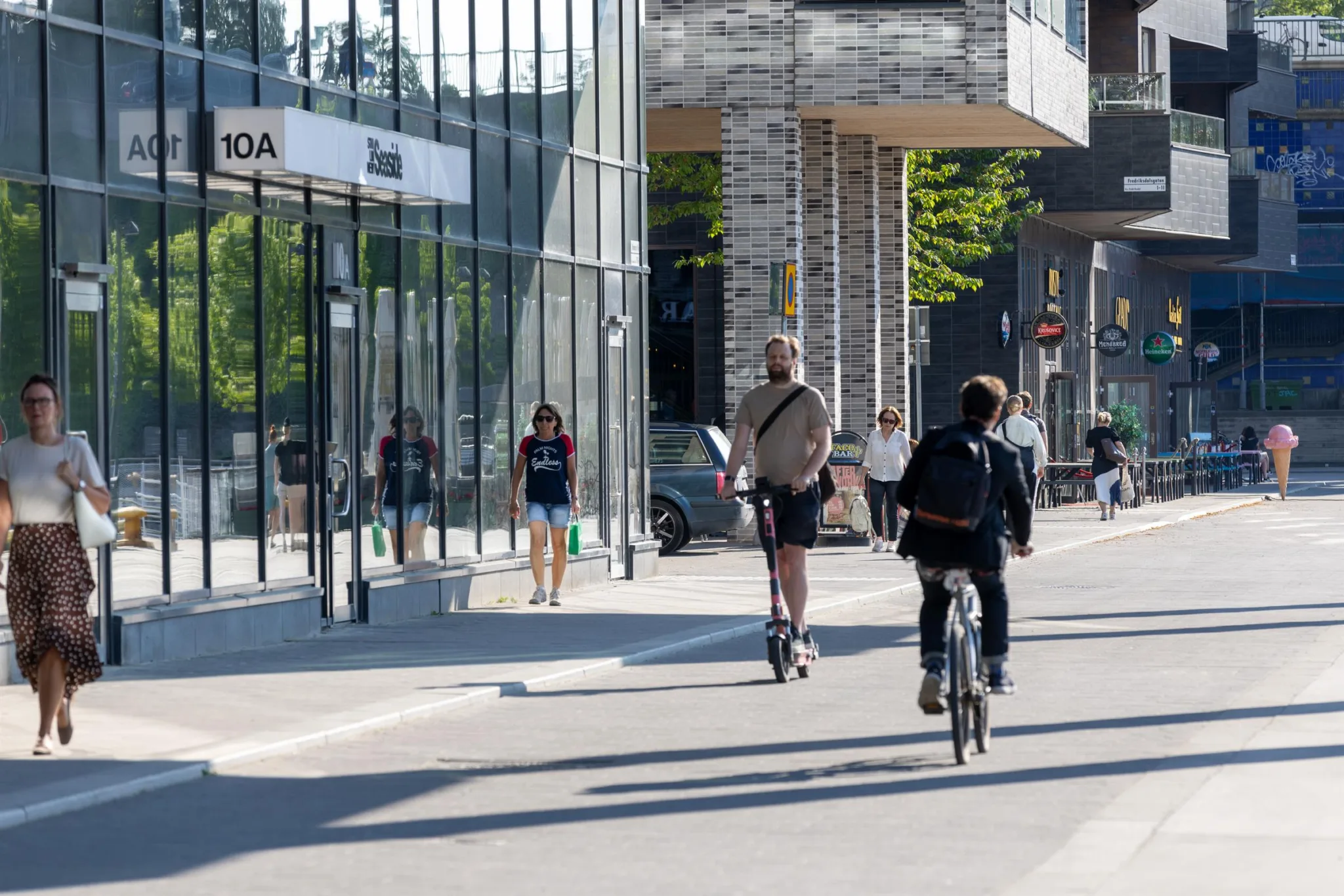 Människor cyklar och åker elsparkcyklar på en gata med moderna byggnader och glasfasader. Trottoaren har flera fotgängare.