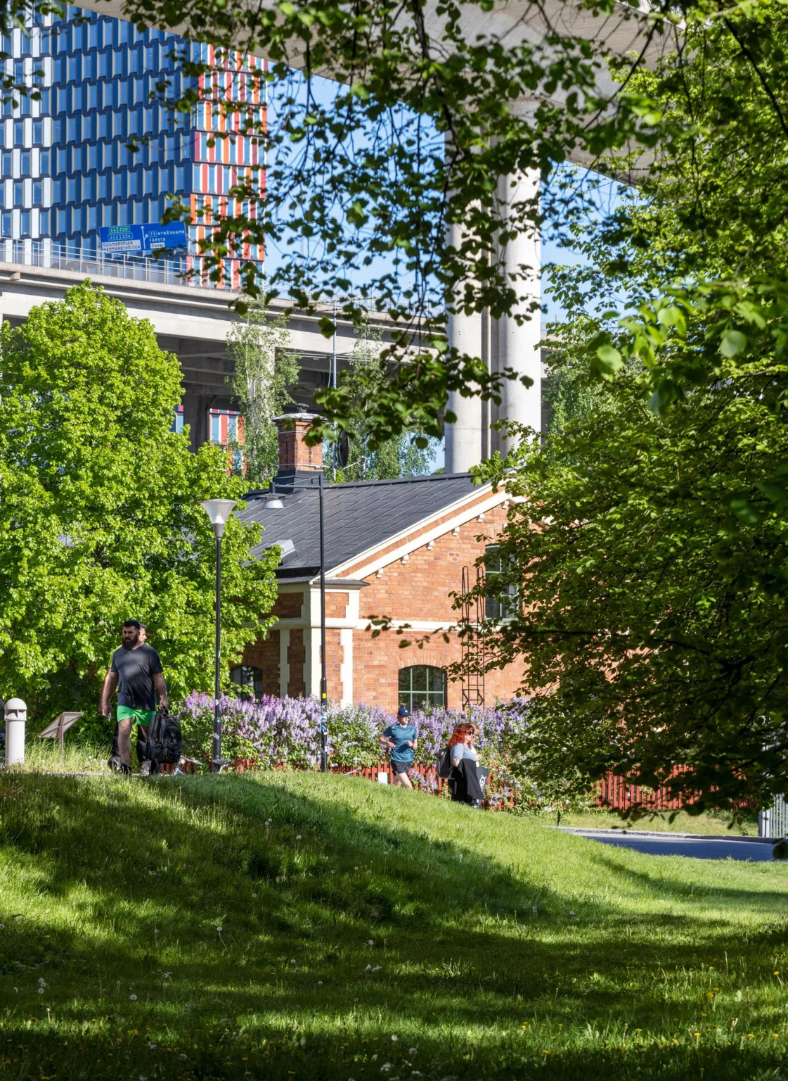 En lummig park med grönska, blommor och en röd tegelbyggnad i bakgrunden.