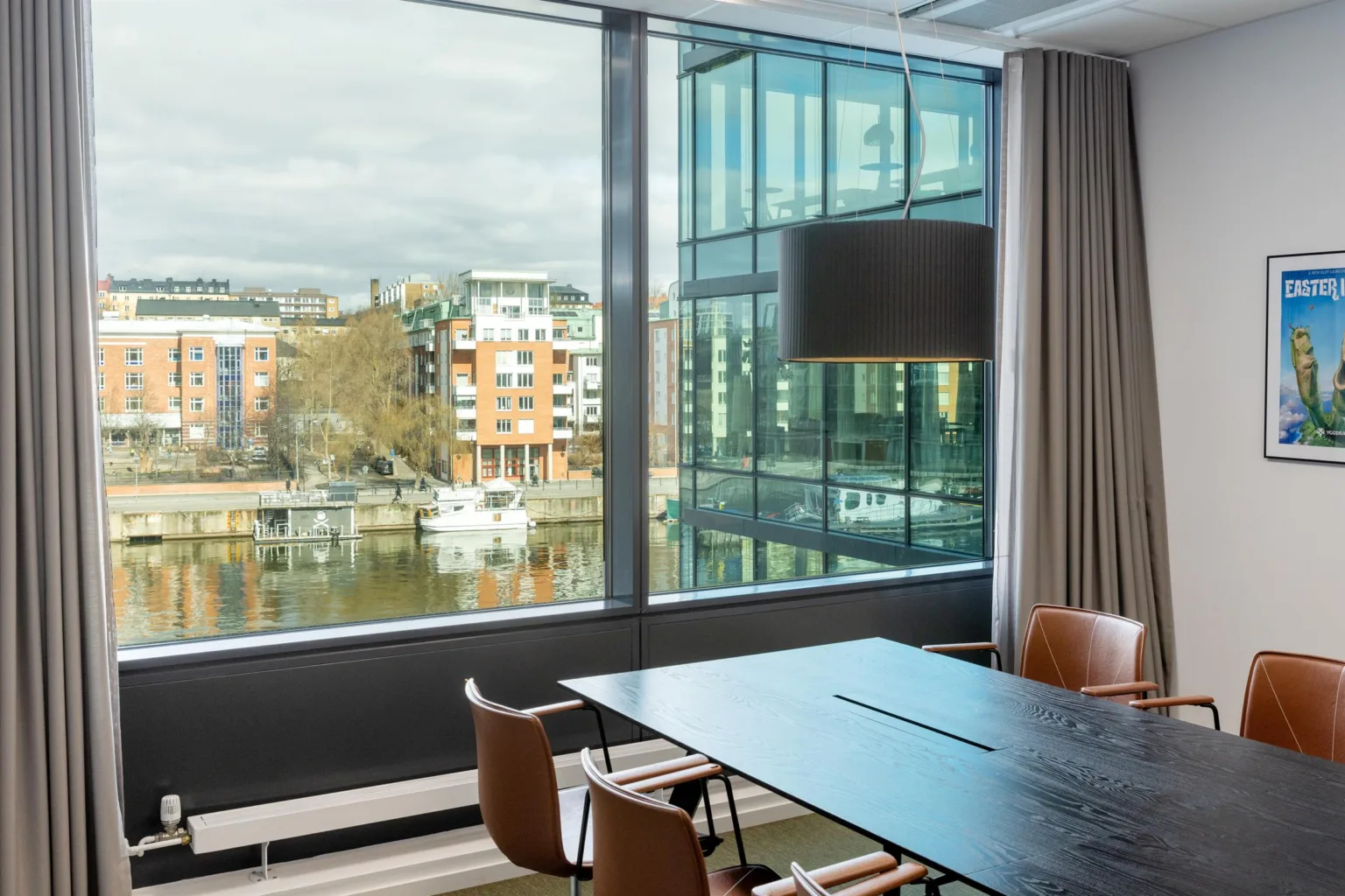 Ett mötesrum med ett stort bord och stolar, med utsikt över en kanal och byggnader genom stora fönster.