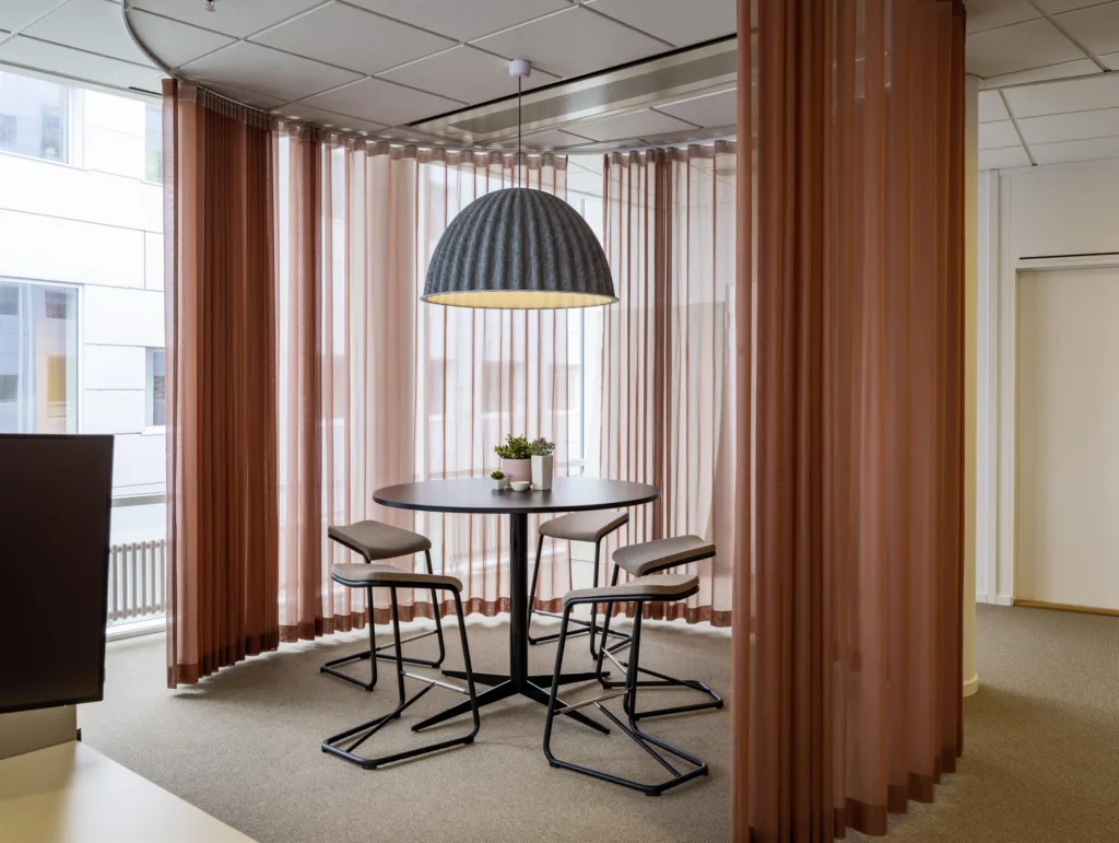 3. Ett modernt mötesrum med ett runt bord, fyra stolar och en stor taklampa. Rummet är omgivet av beige gardiner och glasfönster.