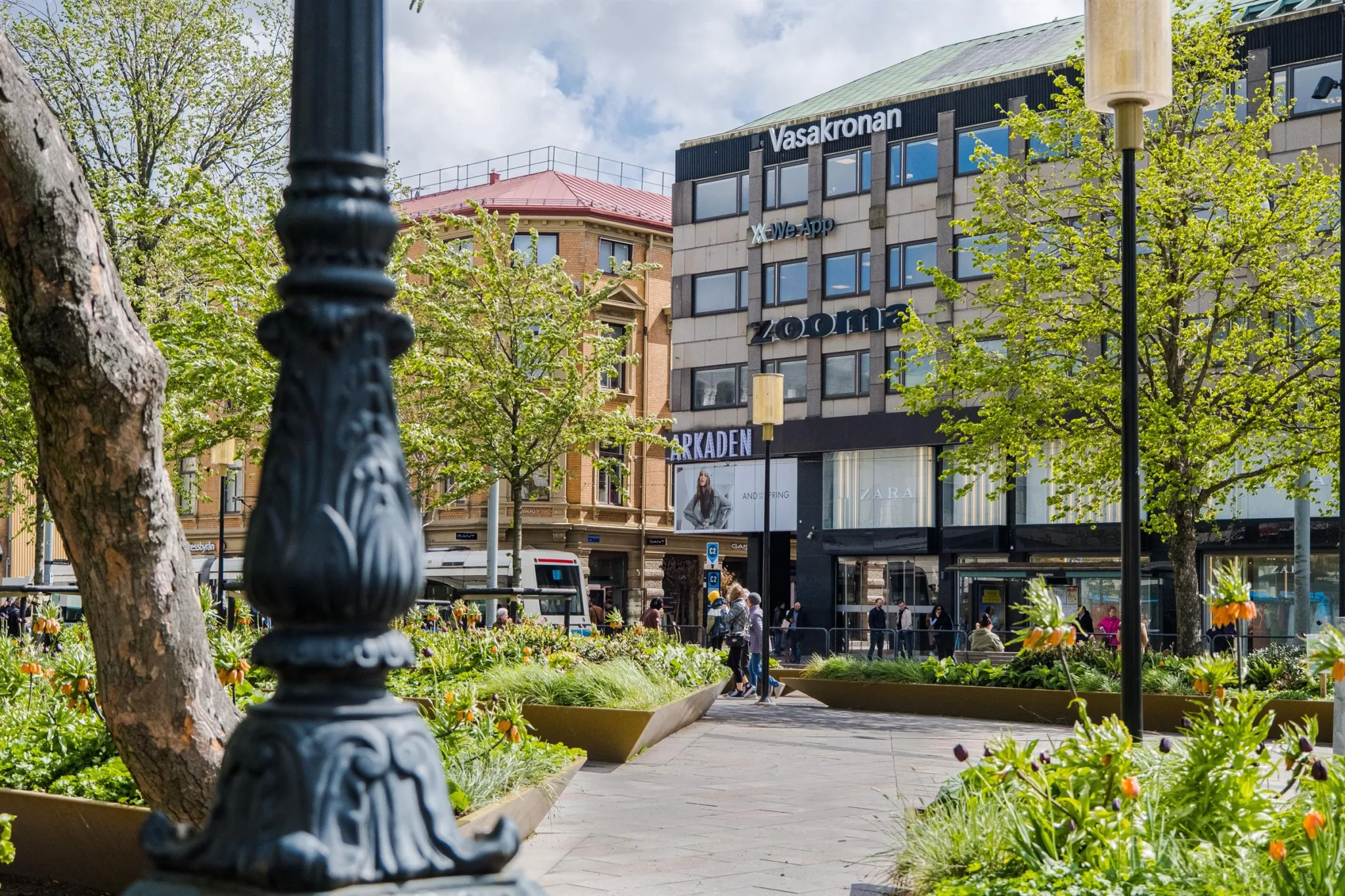 En park med gröna växter, en dekorativ gatlykta i förgrunden och en byggnad med skylten "Zooma" i bakgrunden.