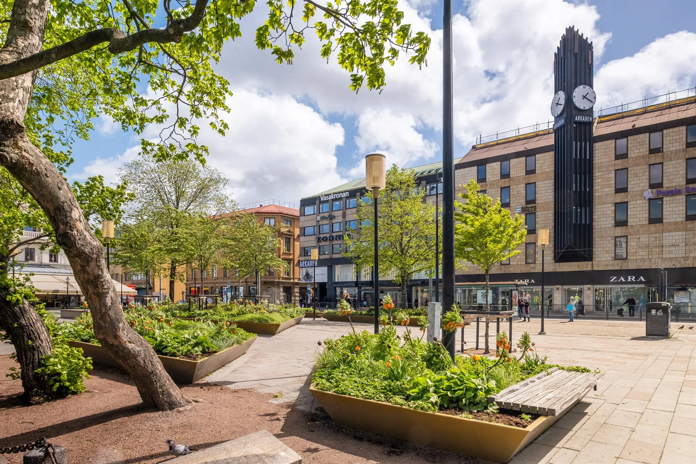 En stadsmiljö med grönskande träd, blomlådor och byggnader under en klarblå himmel.