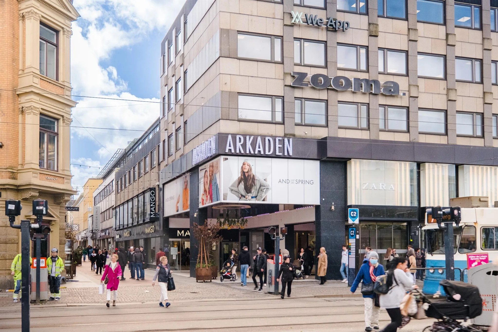Folk går förbi byggnaden "Arkaden" med affärer och reklam under en solig dag.