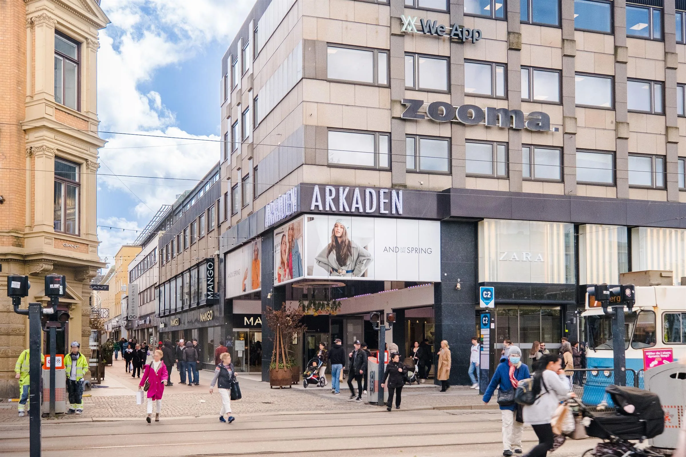 Folk går förbi byggnaden "Arkaden" med affärer och reklam under en solig dag.