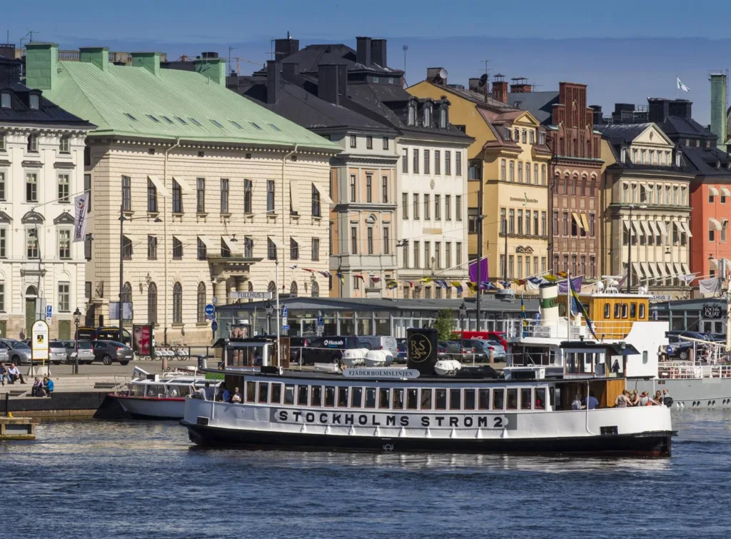 En vit passagerarbåt med texten "Stockholms Ström" vid en kaj med färgglada äldre byggnader.