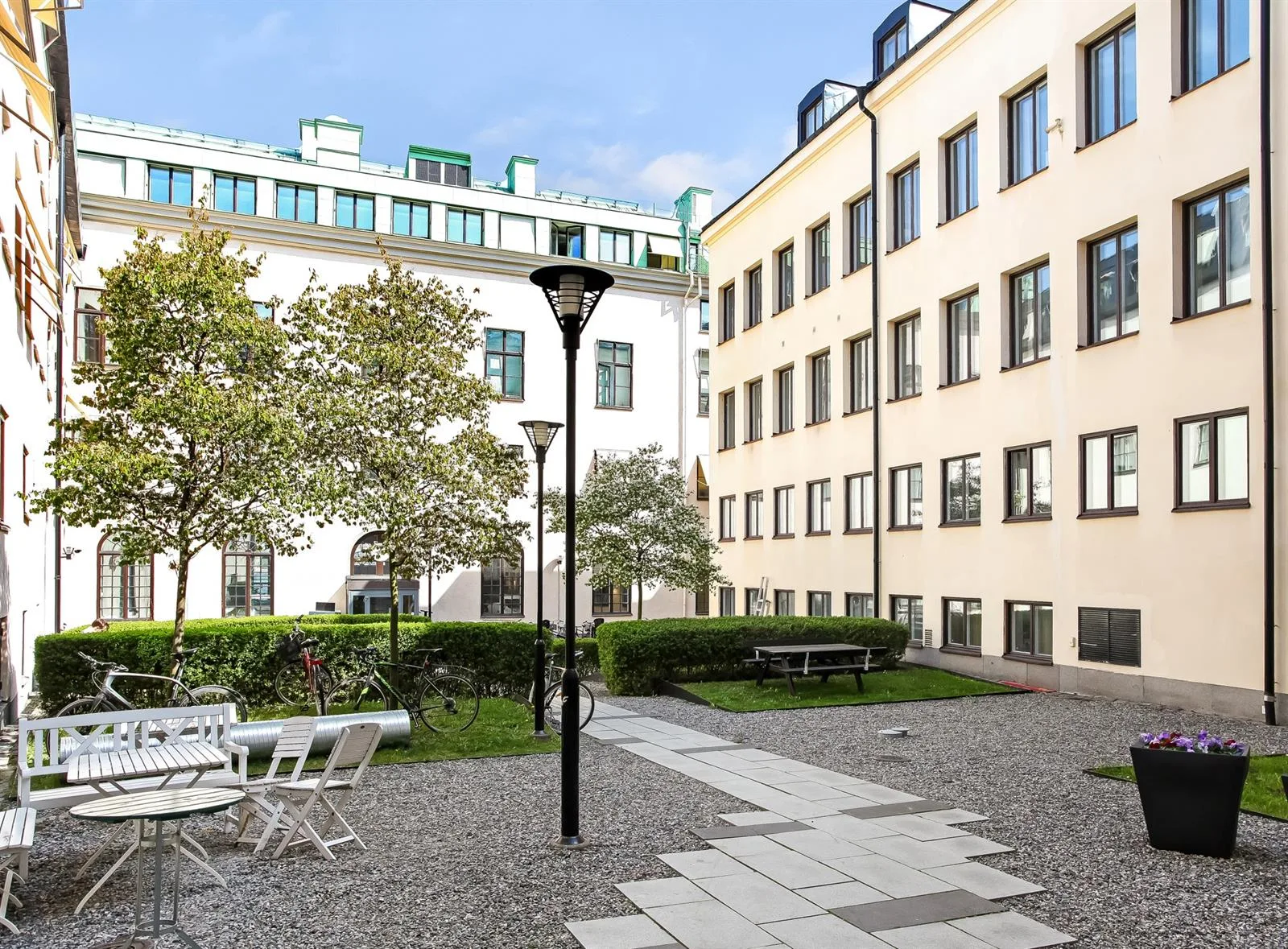 Innergård med grusgångar, bänkar, bord och grönska omgiven av vitmålade byggnader.