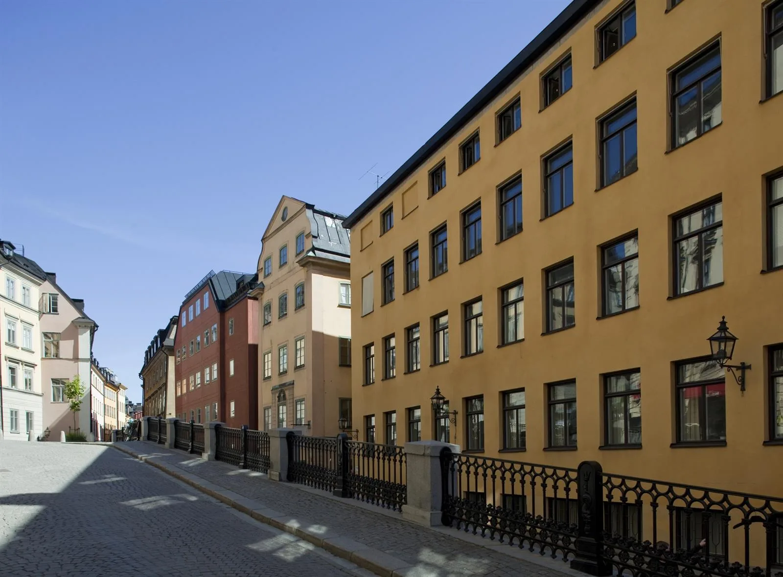 Fasad av gula och röda flerfamiljshus längs en gata med trottoar och staket.