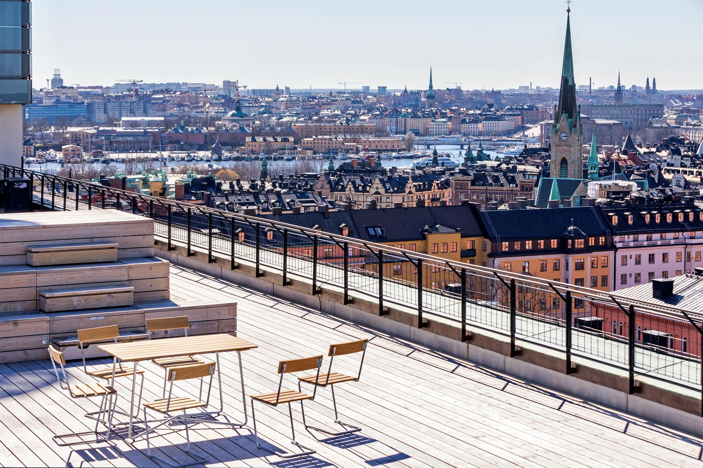En takterrass med bord och stolar med utsikt över Stockholm, inklusive kyrkspiror och hustak.