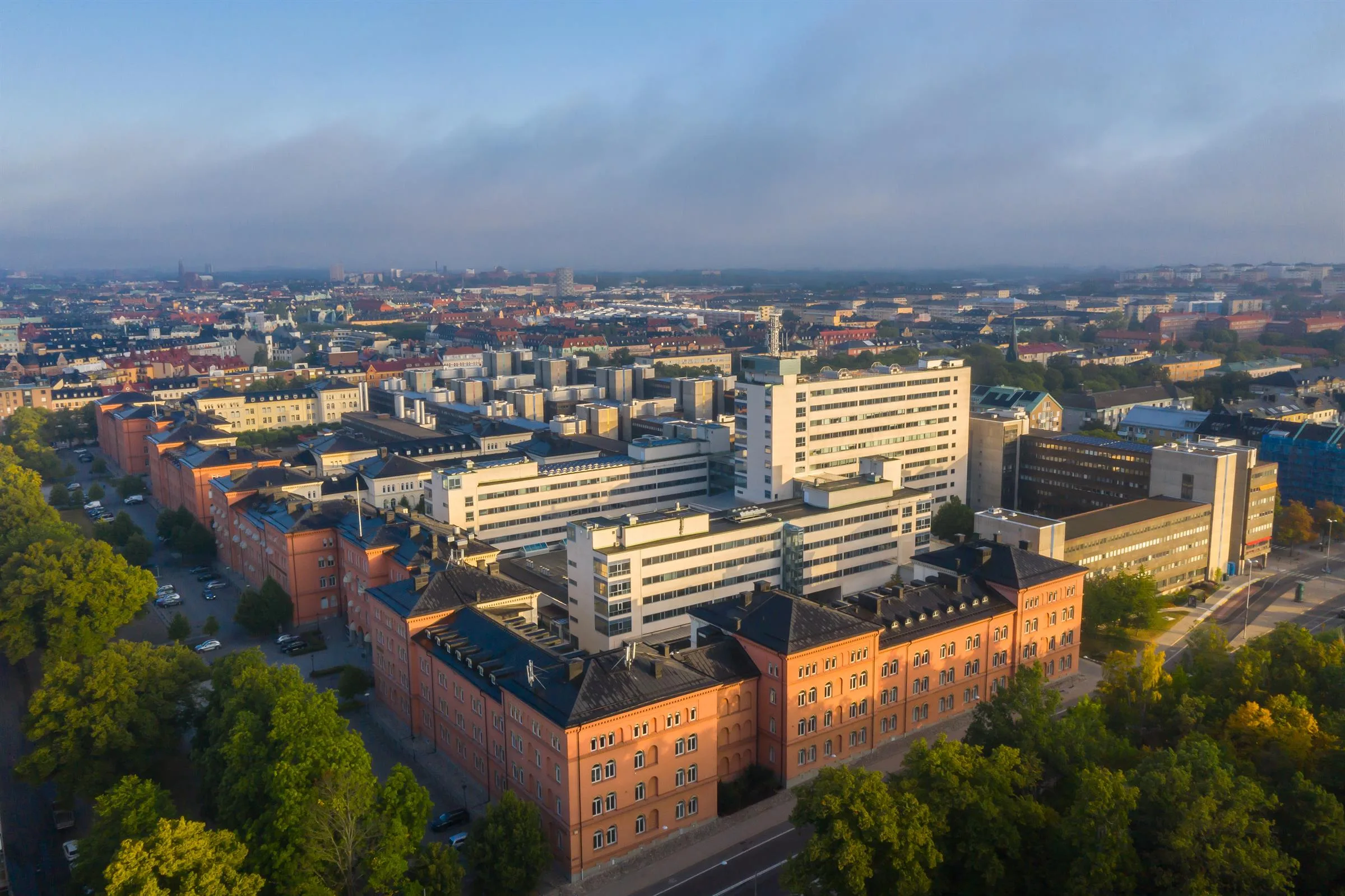 En vy ovanför en stadsdel med vita byggnader och röda tegelhus, omgiven av grönska under en blå himmel.