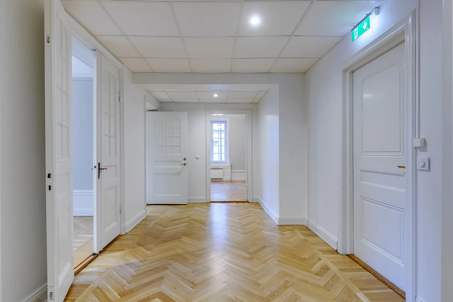 2. En korridor med parkettgolv, vita väggar och flera vita dörrar samt en nödutgångsskylt.