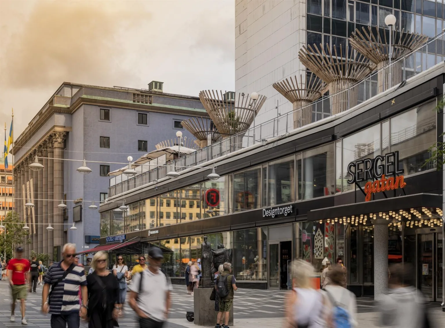En gata med människor som passerar förbi butiker och byggnader, med modern arkitektur och skulpturer ovanför.
