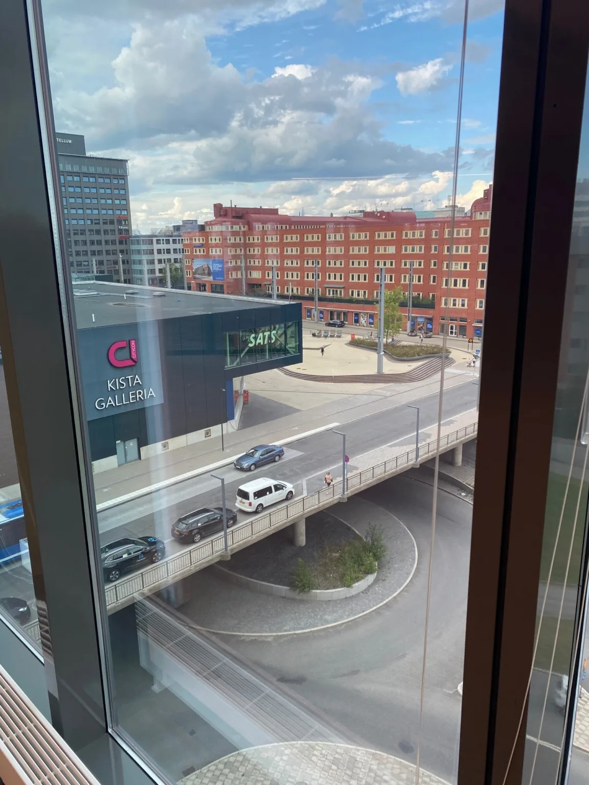 Utsikt från ett fönster mot en stadsmiljö med en väg, byggnader och en blå himmel.