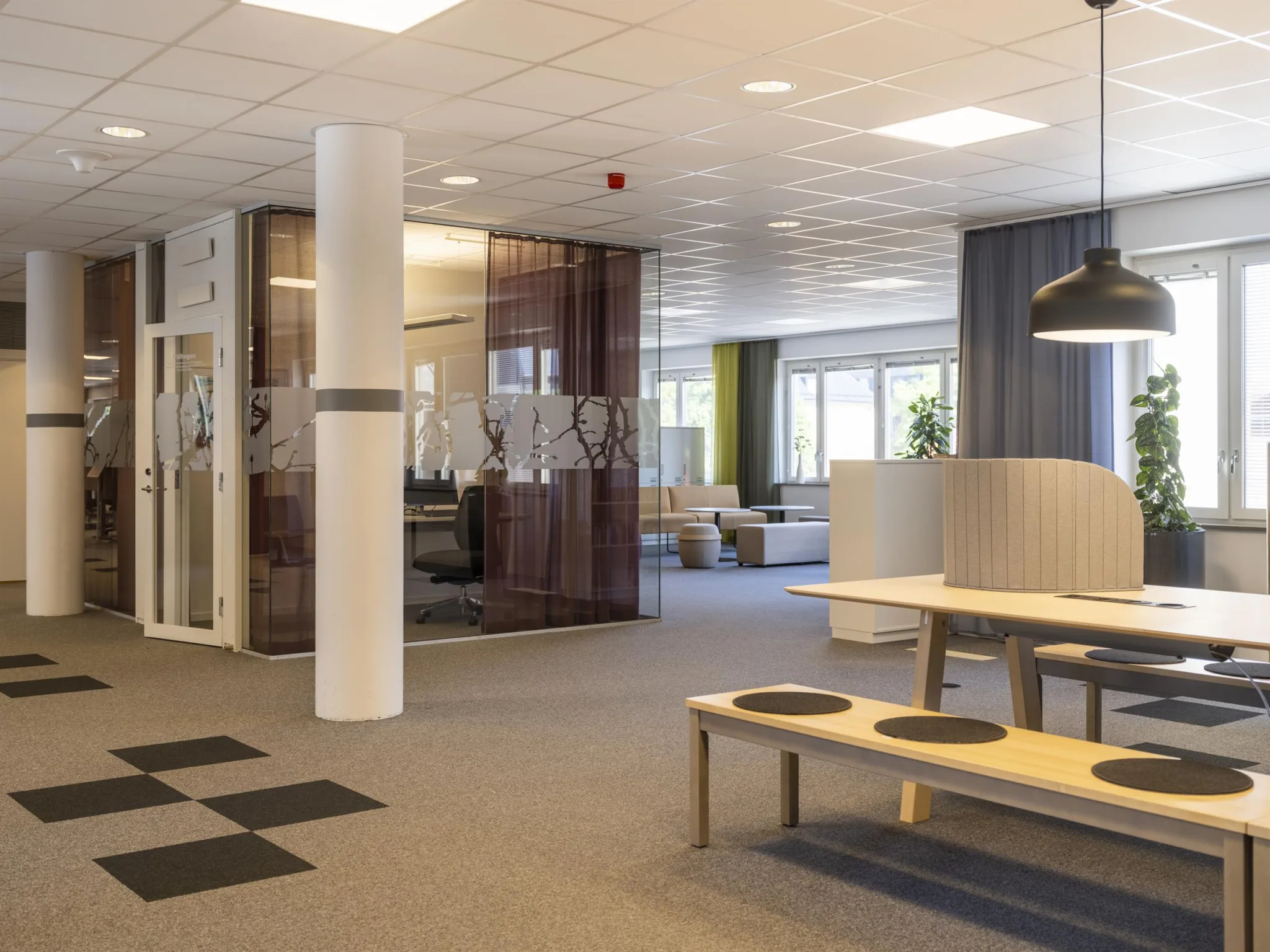 Ett modernt kontorsutrymme med bord, bänkar och glasväggar, samt stora fönster som släpper in naturligt ljus.