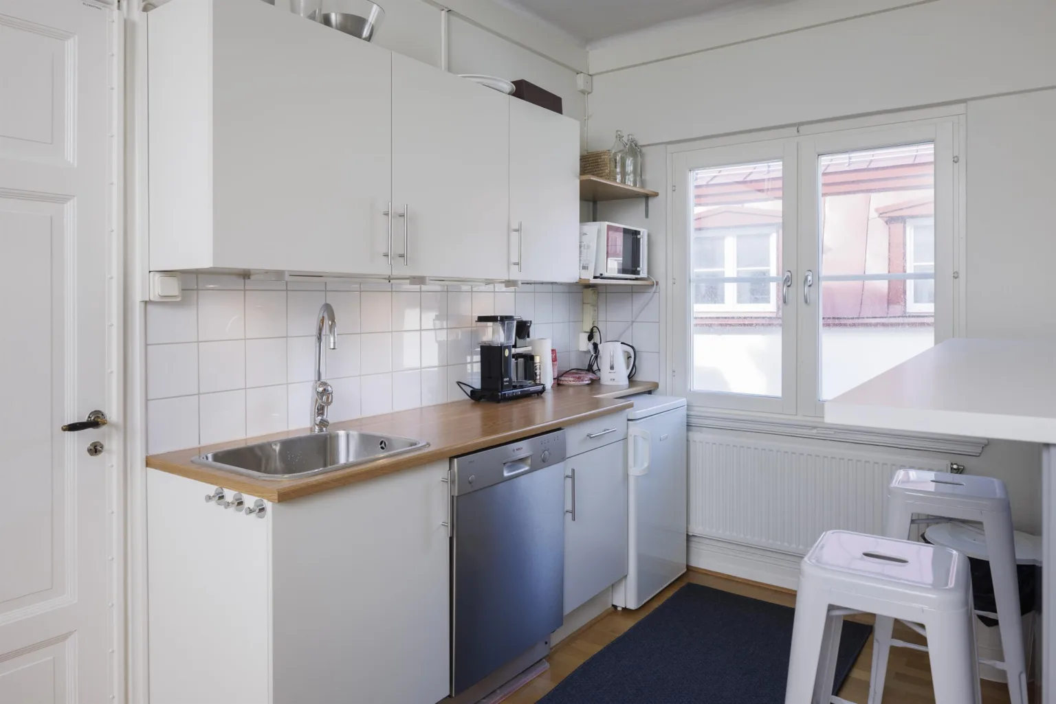 Kök med vita skåp, träbänkskivor, diskho, diskmaskin och kaffemaskin. Fönster med utsikt mot byggnader.