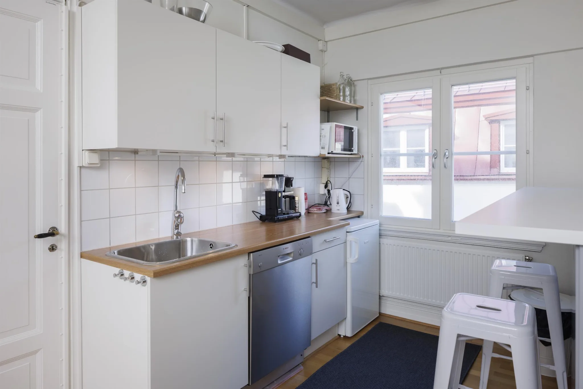 Kök med vita skåp, träbänkskivor, diskho, diskmaskin och kaffemaskin. Fönster med utsikt mot byggnader.