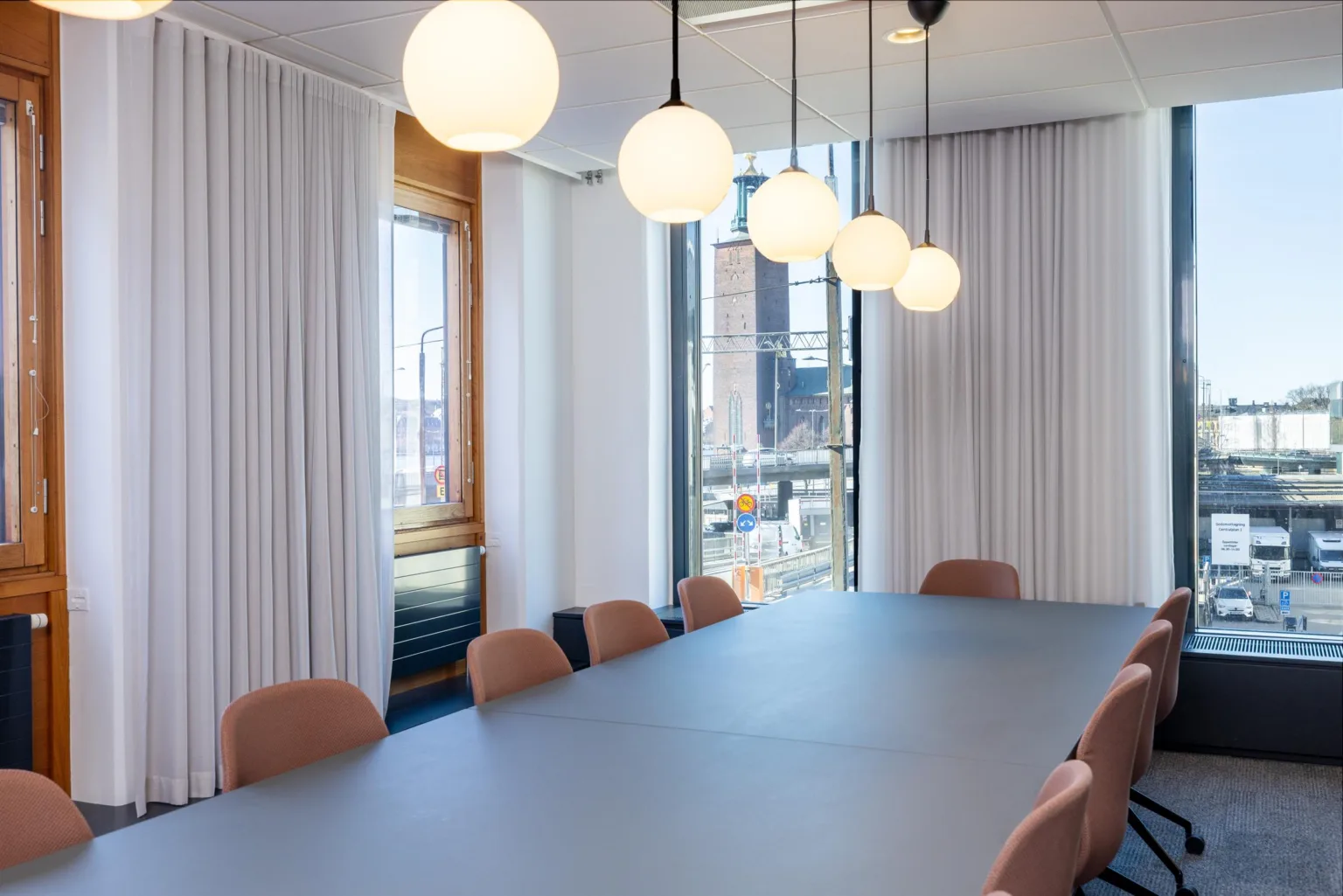 Ett elegant konferensrum med ett stort blått bord, rosa stolar och hängande lampor framför stora fönster täckta av vita gardiner.
