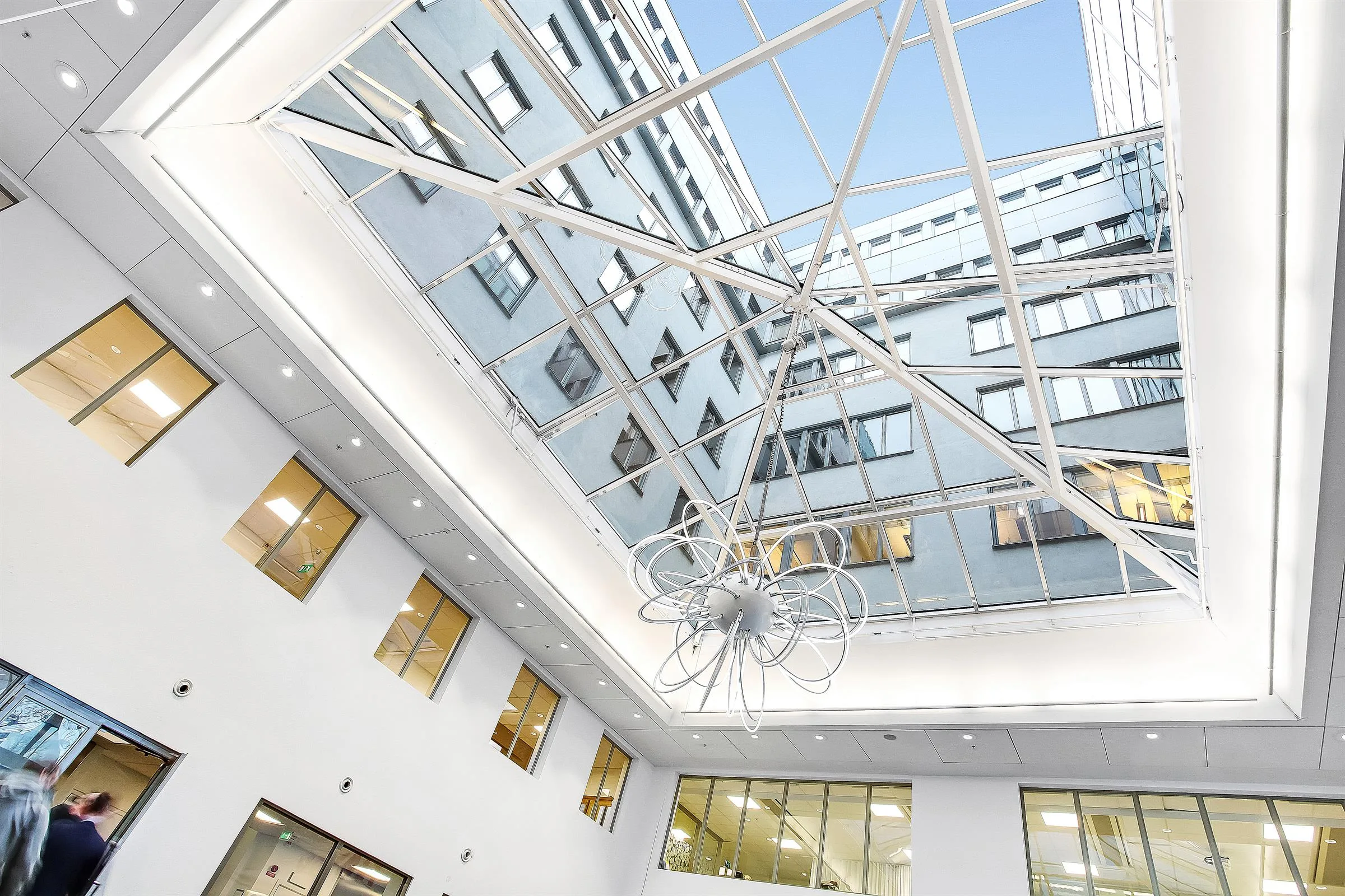 Ett modernt atrium med höga fönster, vit interiör och ett glastak som släpper in dagsljus.