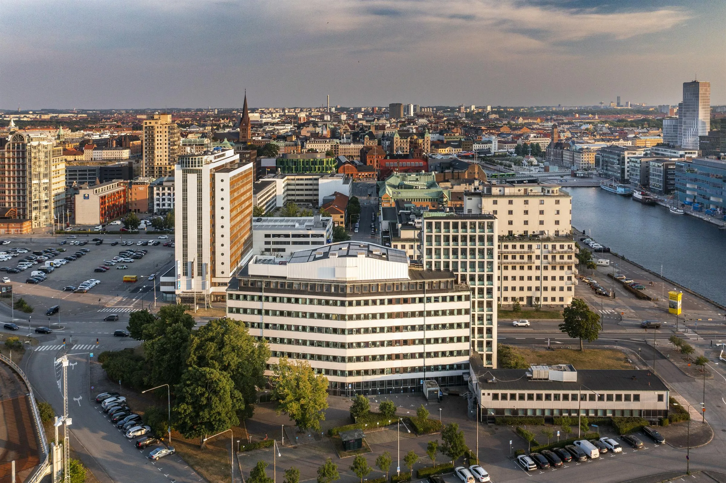 En stadsbild med flera byggnader, inklusive höghus, parkeringsytor och en vattenspegel i bakgrunden.