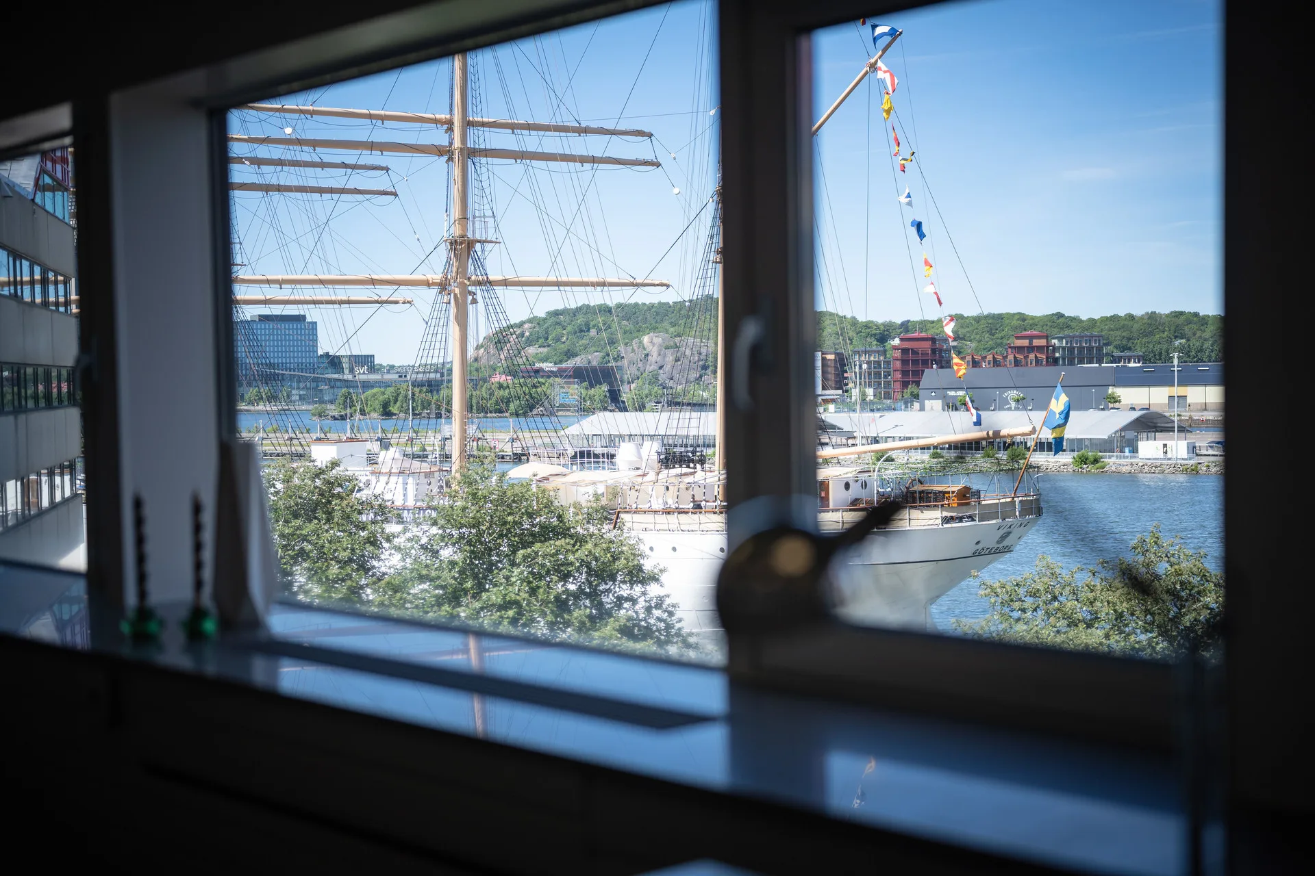 3. Utsikt genom ett fönster mot ett segelfartyg, med grönska och vatten utanför.