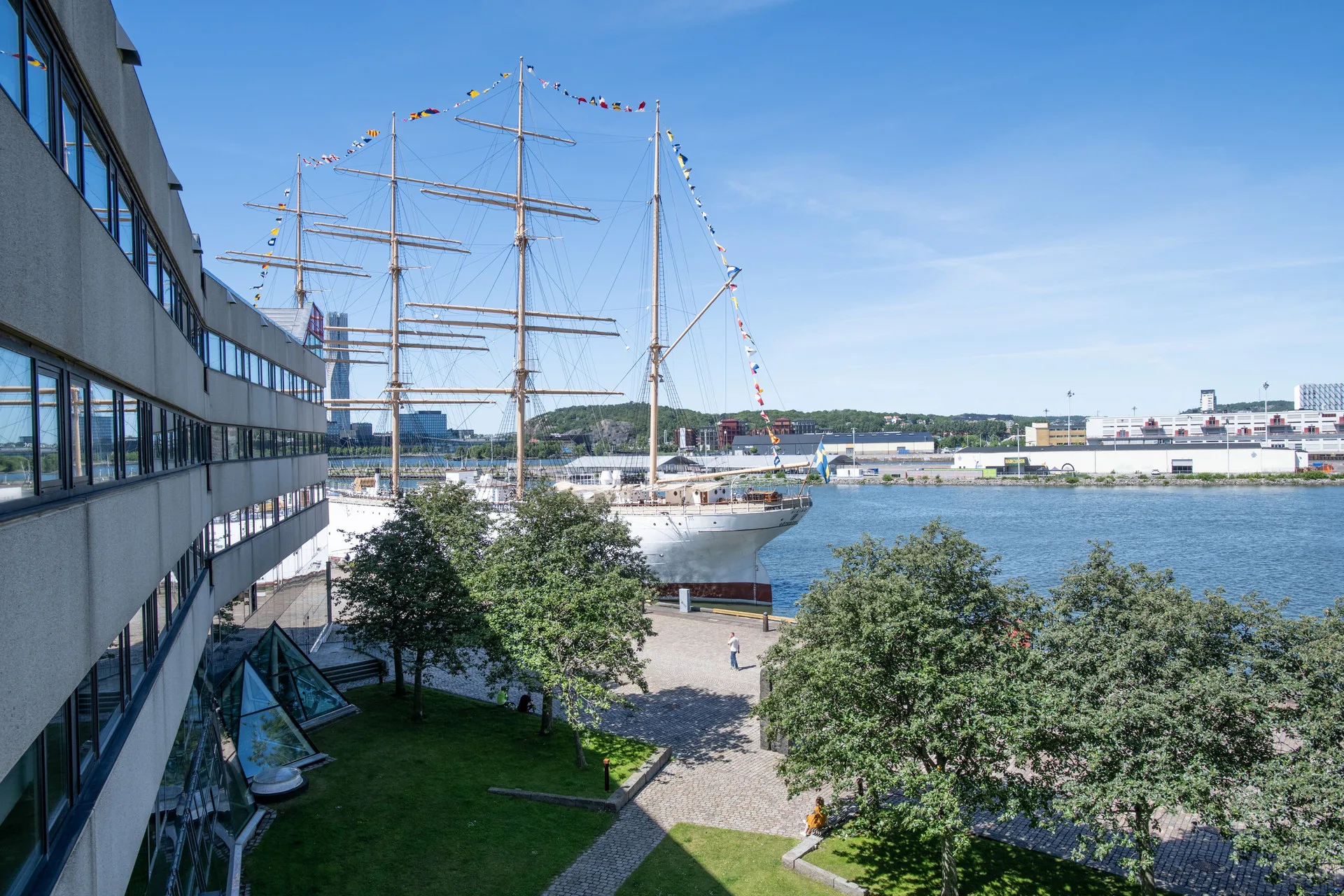 1. Ett stort segelfartyg ligger vid kajen nära ett grönt grönområde, med en byggnad till vänster och vatten i bakgrunden.