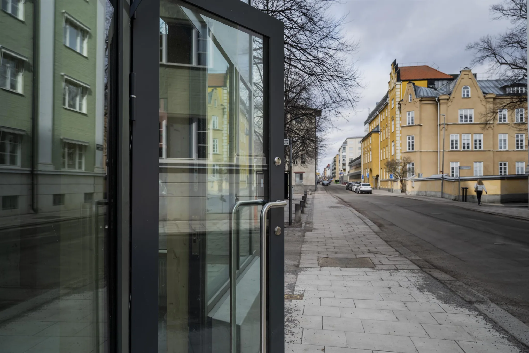 En glasdörr med metallhandtag i förgrunden, med en trottoar och gula byggnader längs en gata i bakgrunden.