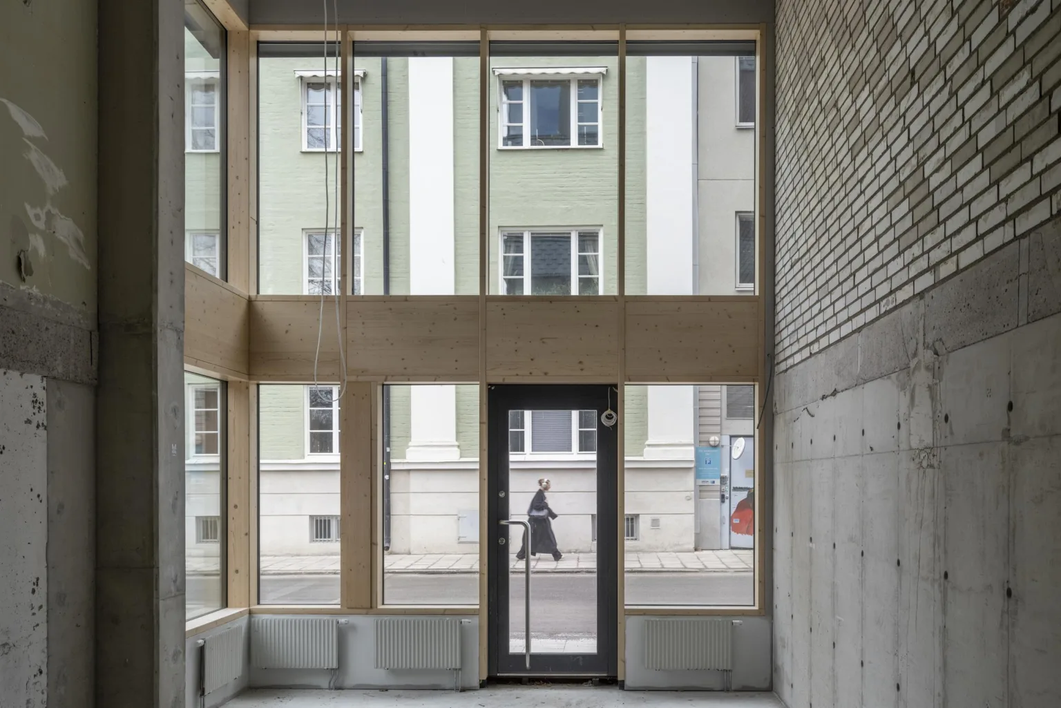 En glasentré med stora fönster, med utsikt mot en grön byggnad och en person som går på gatan utanför.