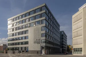 En modern kontorsbyggnad med stora glasfönster och geometrisk design, belägen vid en gata med blå himmel ovanför.