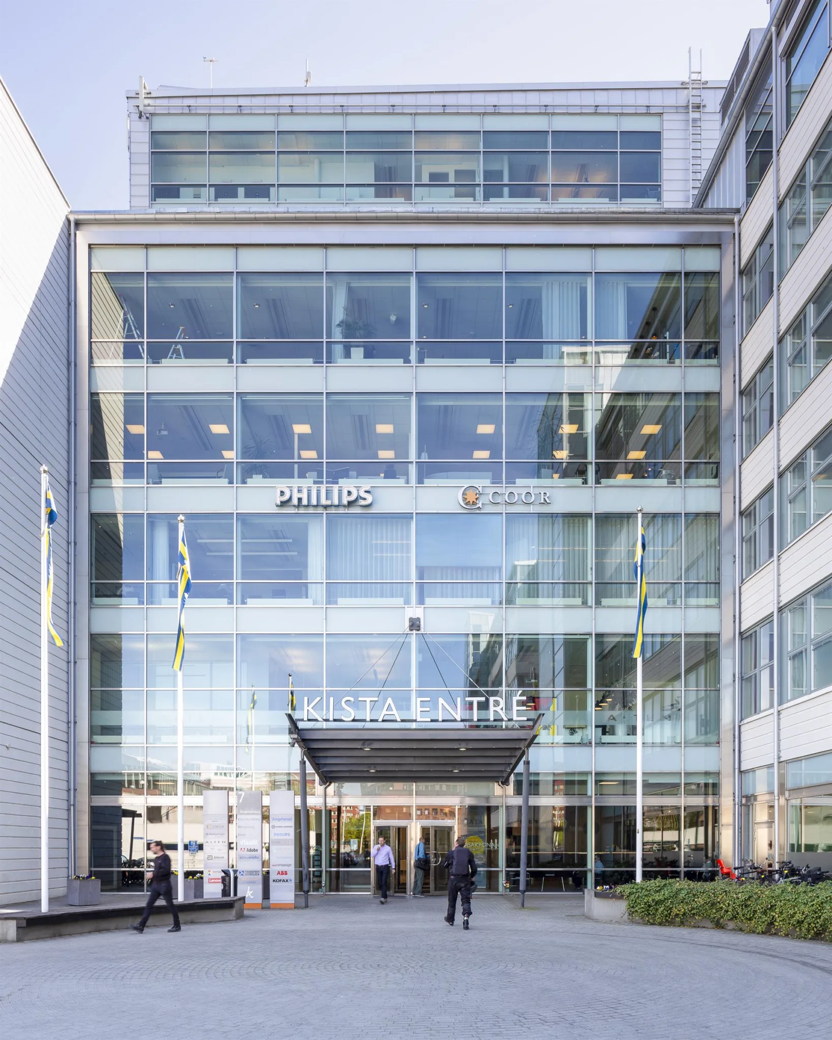 3. Glasfasad på en byggnad med flaggor och texten "KTH Entré" ovanför ingången, samt personer som går in och ut.