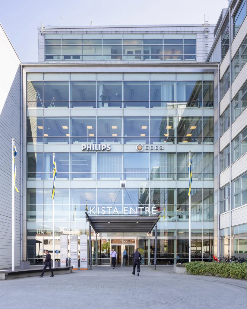 3. Glasfasad på en byggnad med flaggor och texten "KTH Entré" ovanför ingången, samt personer som går in och ut.