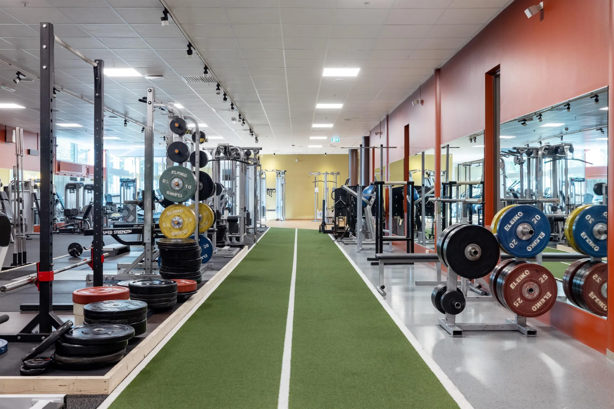 Inomhusgym med träningsutrustning och en grön löparbana i mitten av rummet.