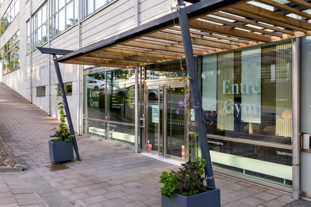 5. Ingång till en byggnad med skylttexten "Entré Gym" på glasdörren, omgiven av blomkrukor och en modern överbyggnad.