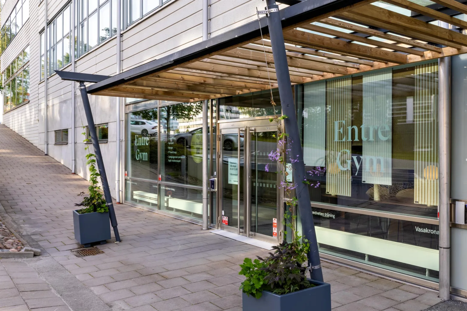 5. Ingång till en byggnad med skylttexten "Entré Gym" på glasdörren, omgiven av blomkrukor och en modern överbyggnad.