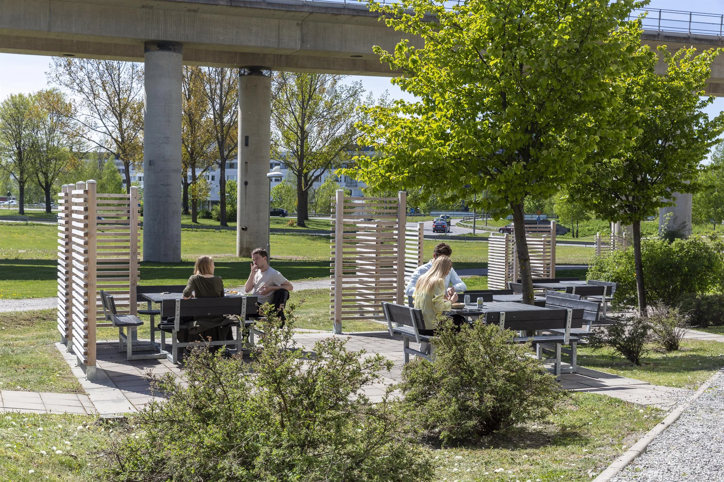 Personer sitter vid utomhusbord i en grön park med träd och buskar, samt en gångbro i bakgrunden.
